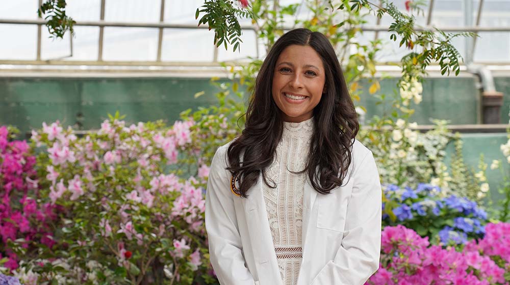 Claire Ginsberg, wearing a white dress and coat, stands in front of blooming flower bushes and smiles.